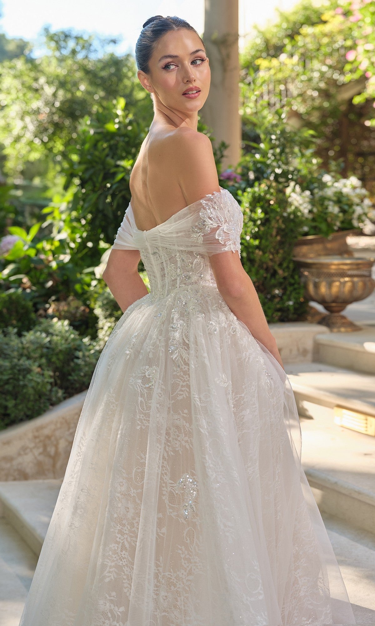 A woman in the Andrea and Leo WL090 long wedding dress, featuring an off-the-shoulder lace design with a delicate corset bodice, stands on outdoor steps surrounded by lush greenery and sunlight, turning slightly to look over her shoulder.
